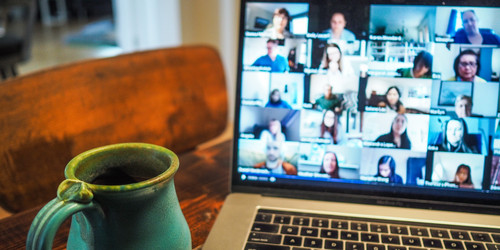 Eine Tasse Kaffe steht an der Seite eines Laptops. Auf dem Monitor sieht man viele Personen, die sich in einer Videokonferenz befinden.