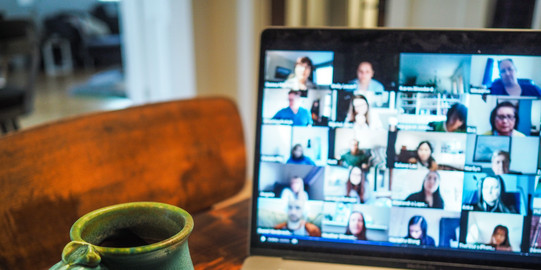 Eine Tasse Kaffe steht an der Seite eines Laptops. Auf dem Monitor sieht man viele Personen, die sich in einer Videokonferenz befinden.