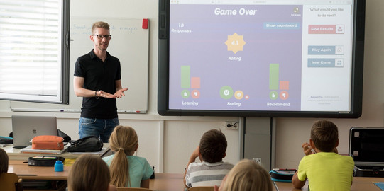 Ein Lehrer steh mit einem Smartboard vor einer Klasse Schüler*innen