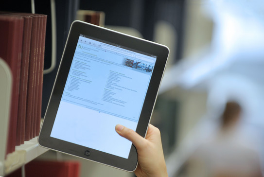 Eine Hand hält ein Tablet in der Universitätsbibliothek an einem Bücherregal.