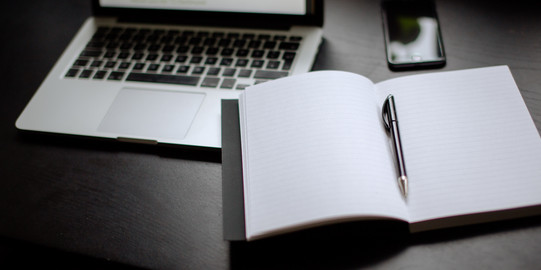 Eingeschalteter Laptop, Smartphone und Notizbuch mit Stift auf einem Tisch liegend.