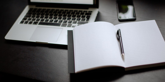 Eingeschalteter Laptop, Smartphone und Notizbuch mit Stift auf einem Tisch liegend.