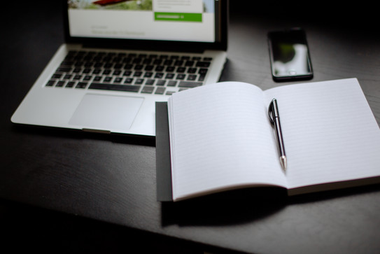 Eingeschalteter Laptop, Smartphone und Notizbuch mit Stift auf einem Tisch liegend.