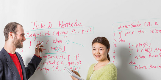 Zwei Personen stehen vor einem Whiteboard mit Formeln und Beschriftung. Eine Person hat einen Stift in der Hand, die andere einen Block.